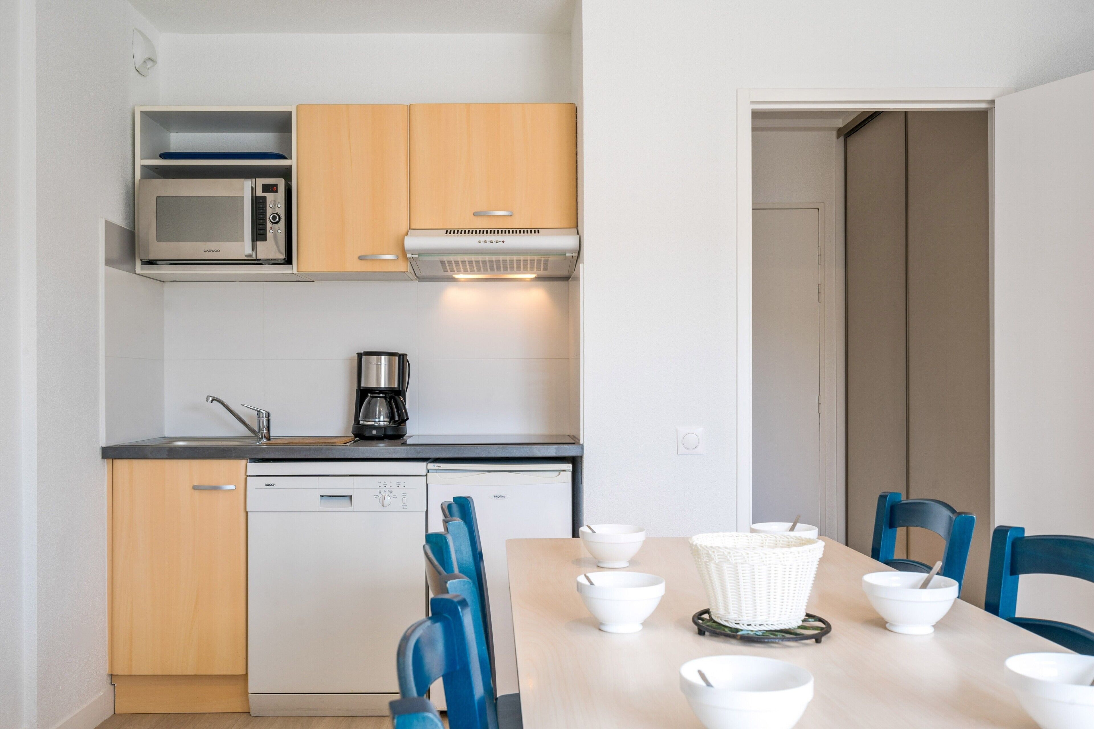 A compact kitchen with a microwave, coffee maker, and dishwasher. There is a dining table with white bowls and a basket.
