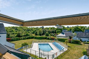A swimming pool with blue lounge chairs, surrounded by a fenced area and a view of a green landscape with houses. - Résidence Pierre et Vacances La Voile d’Or (Île-aux-Moines)