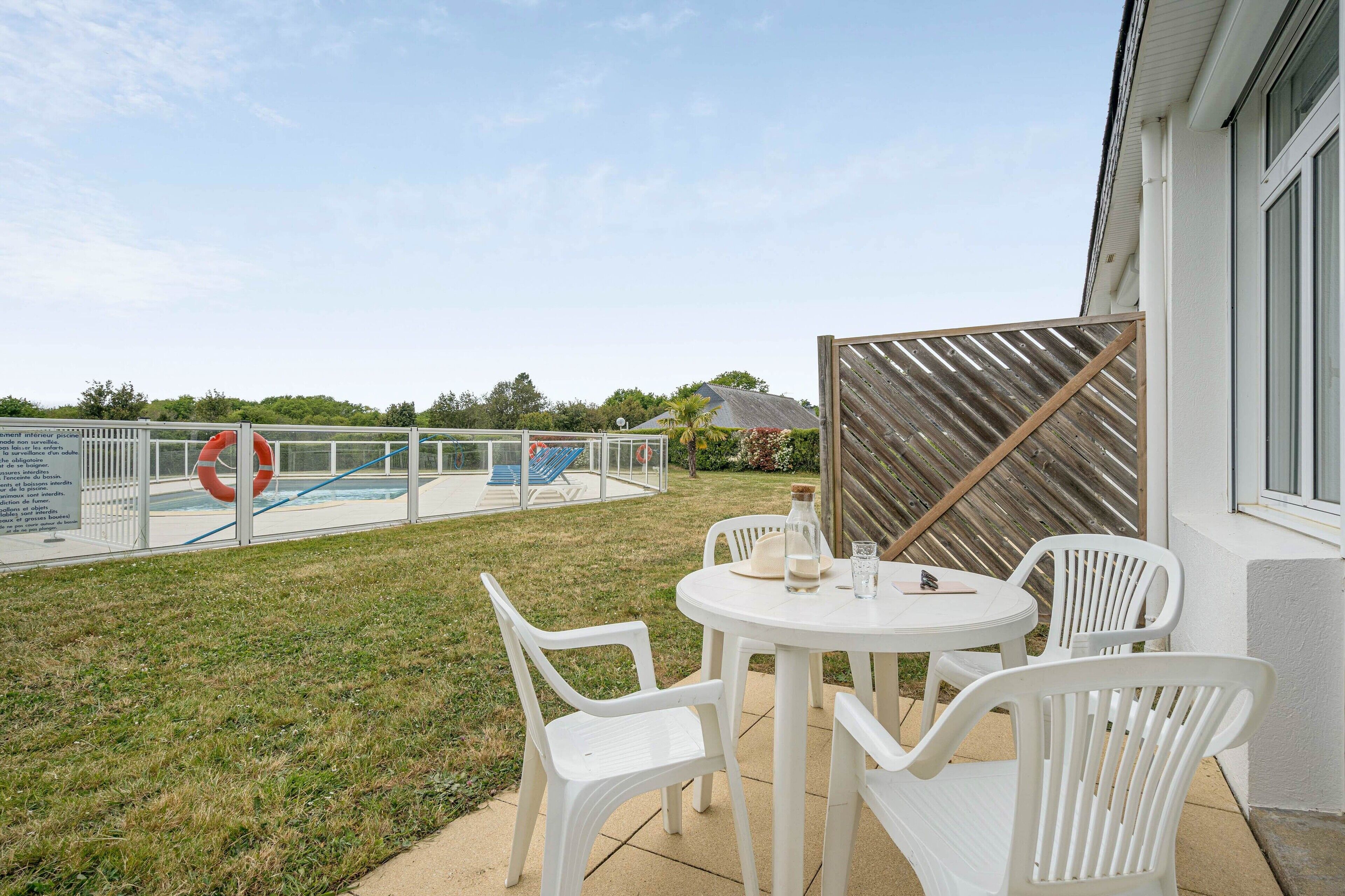 Un patio con sillas de plástico blanco y una mesa, una piscina con un flotador salvavidas y una valla de madera.