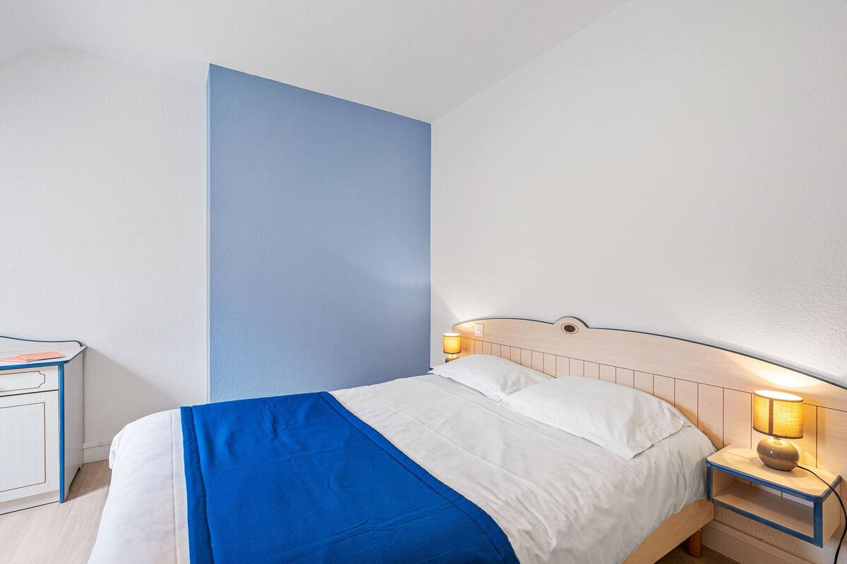 Un lit bien fait, recouvert d’une couvre-lit bleue et blanche, une table de chevet avec une lampe et une armoire blanche.