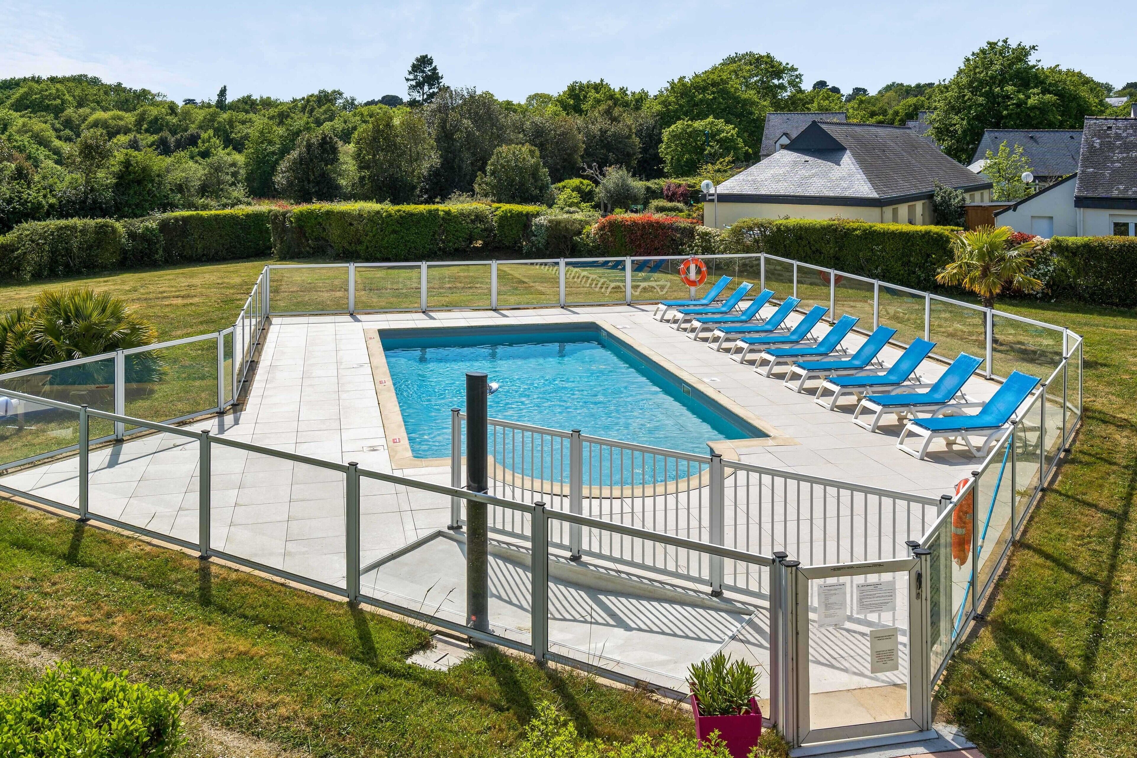 Ein rechteckiger Pool mit einer gefliesten Liegefläche, umgeben von einem Metallzaun. Entlang des Poolrandes befinden sich mehrere blaue Liegestühle.
