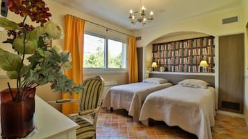 A hotel room with two beds, a bookshelf, a desk with a vase of flowers, and a chair.