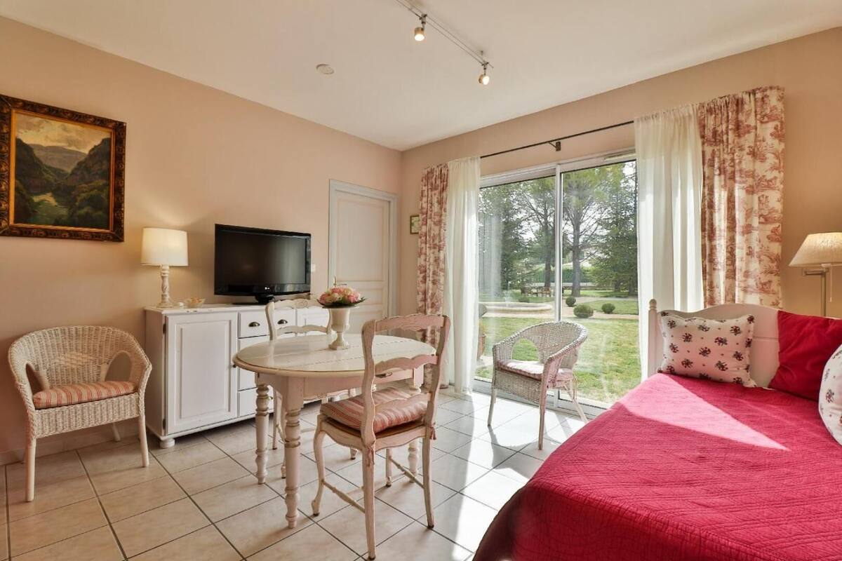 A bedroom with a bed, a TV, a dining table with chairs, and a view of a garden through a sliding door.