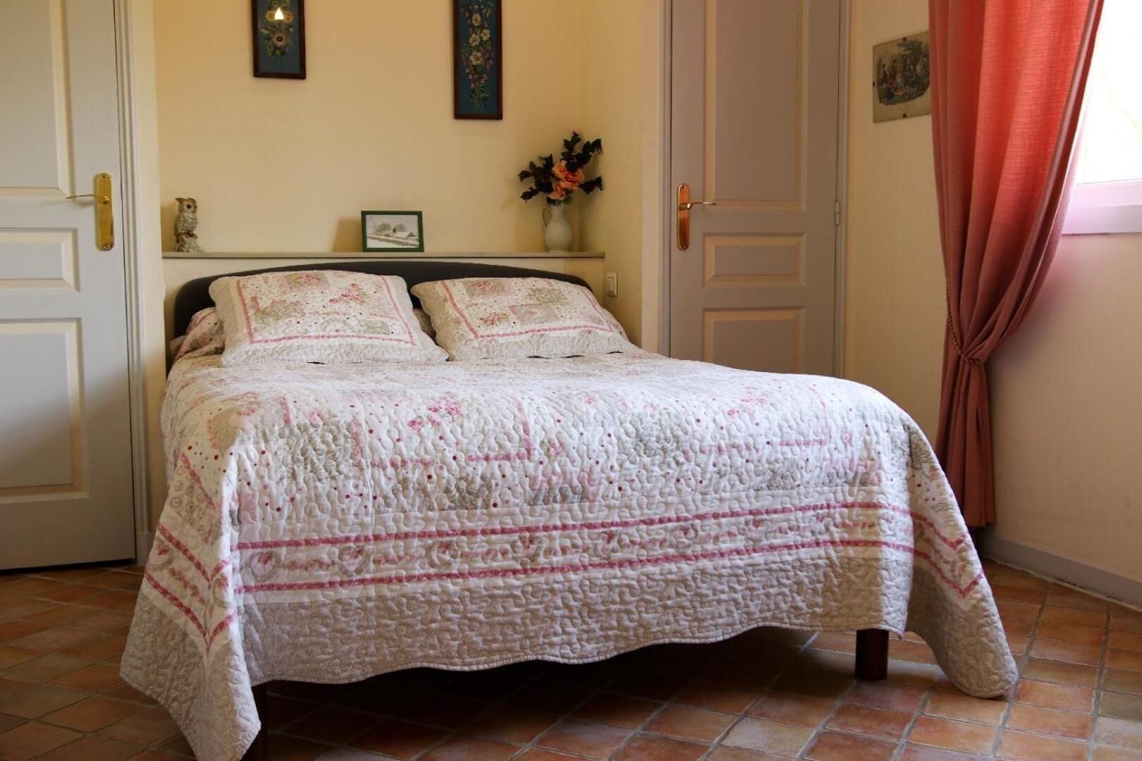 Una cama bien hecha con una colcha estampada y almohadas, una mesita de noche con un jarrón de flores y una puerta con cortinas.
