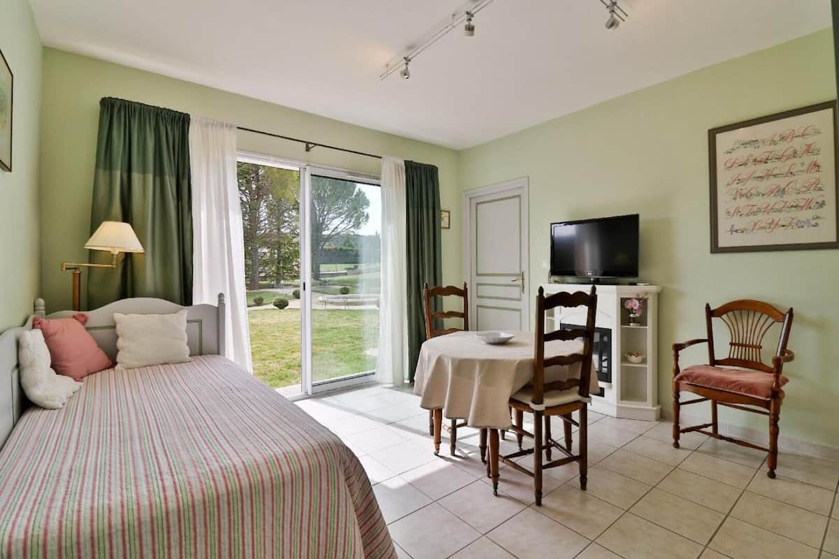 A bedroom with a bed, a table with chairs, a television, and a sliding glass door leading to a garden.