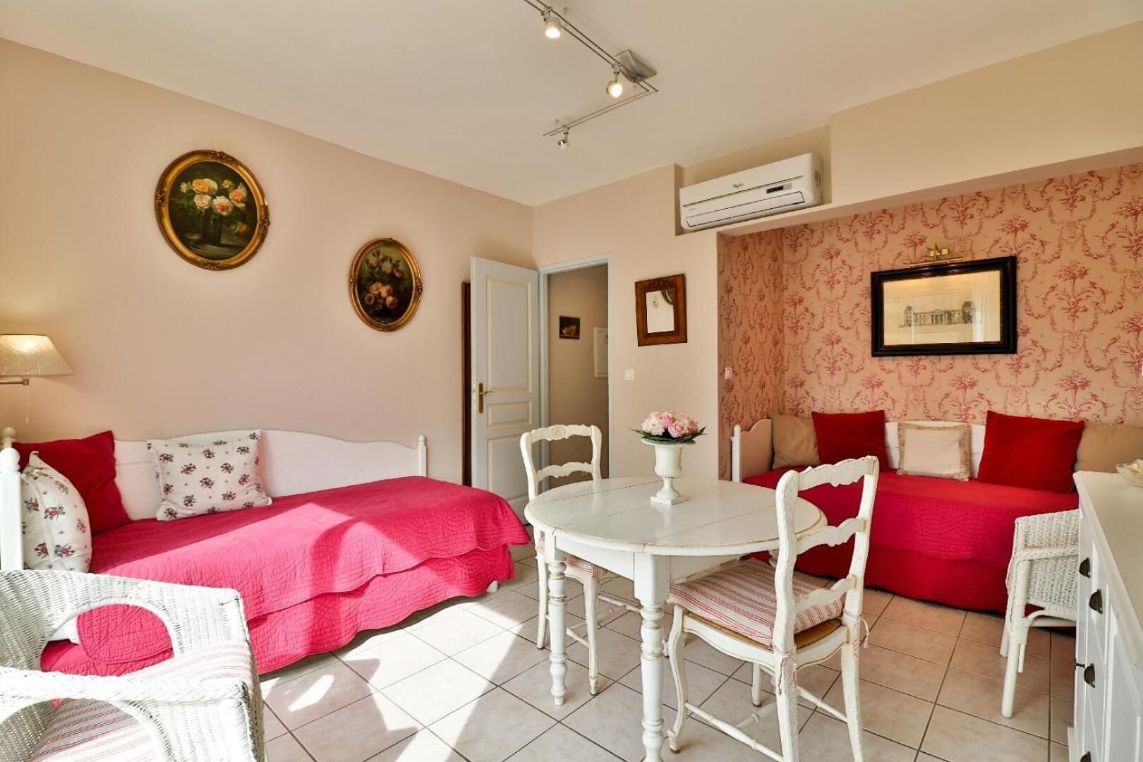 A cozy room with a bed, two chairs, a small table, and a sofa. There are two framed pictures on the wall and an air conditioning unit above the sofa.