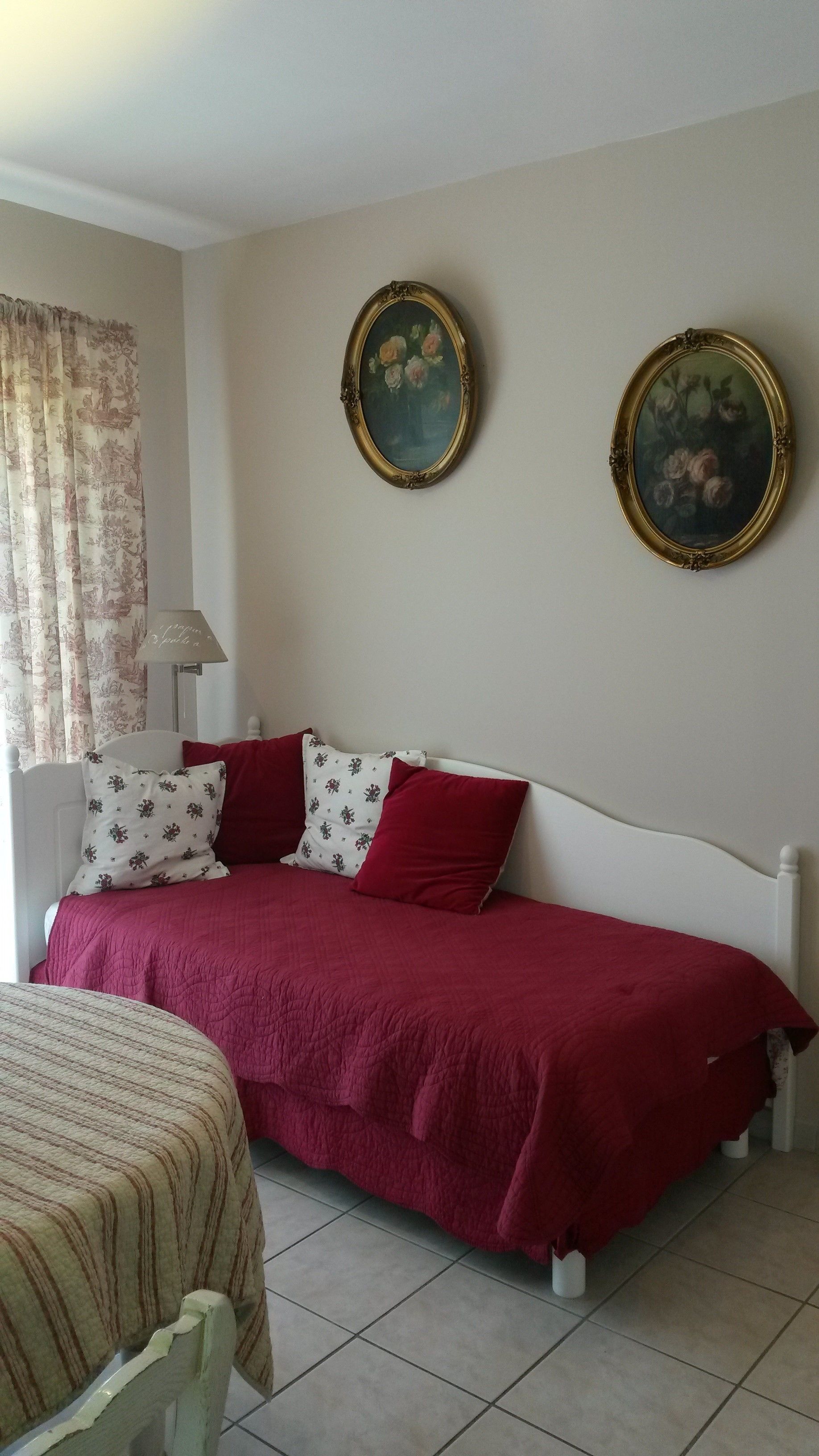Un dormitorio con una cama, dos almohadas, una colcha roja, una lámpara y dos cuadros en la pared.