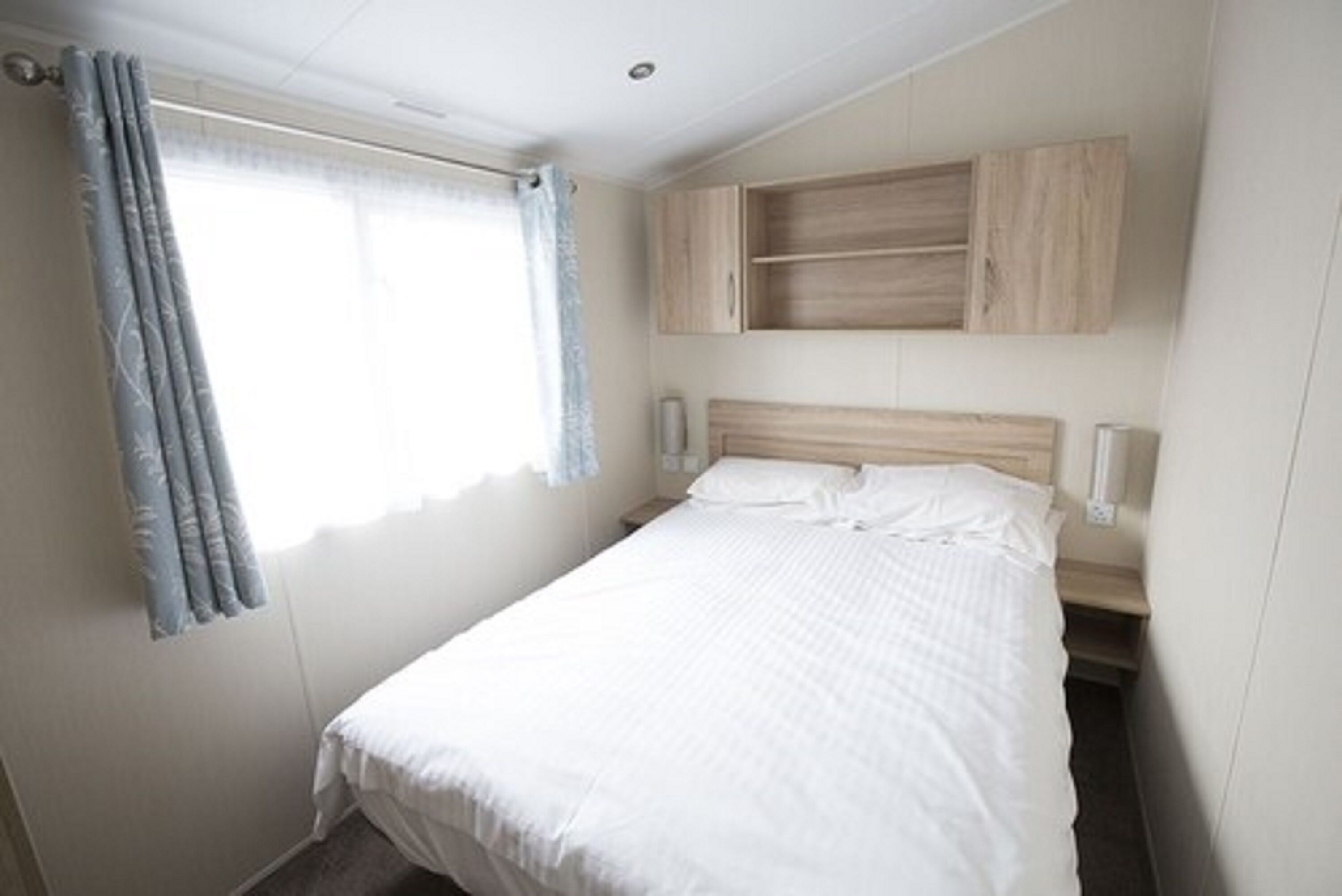 A single bed with a wooden headboard, a window with curtains, and a wooden cabinet above the bed.