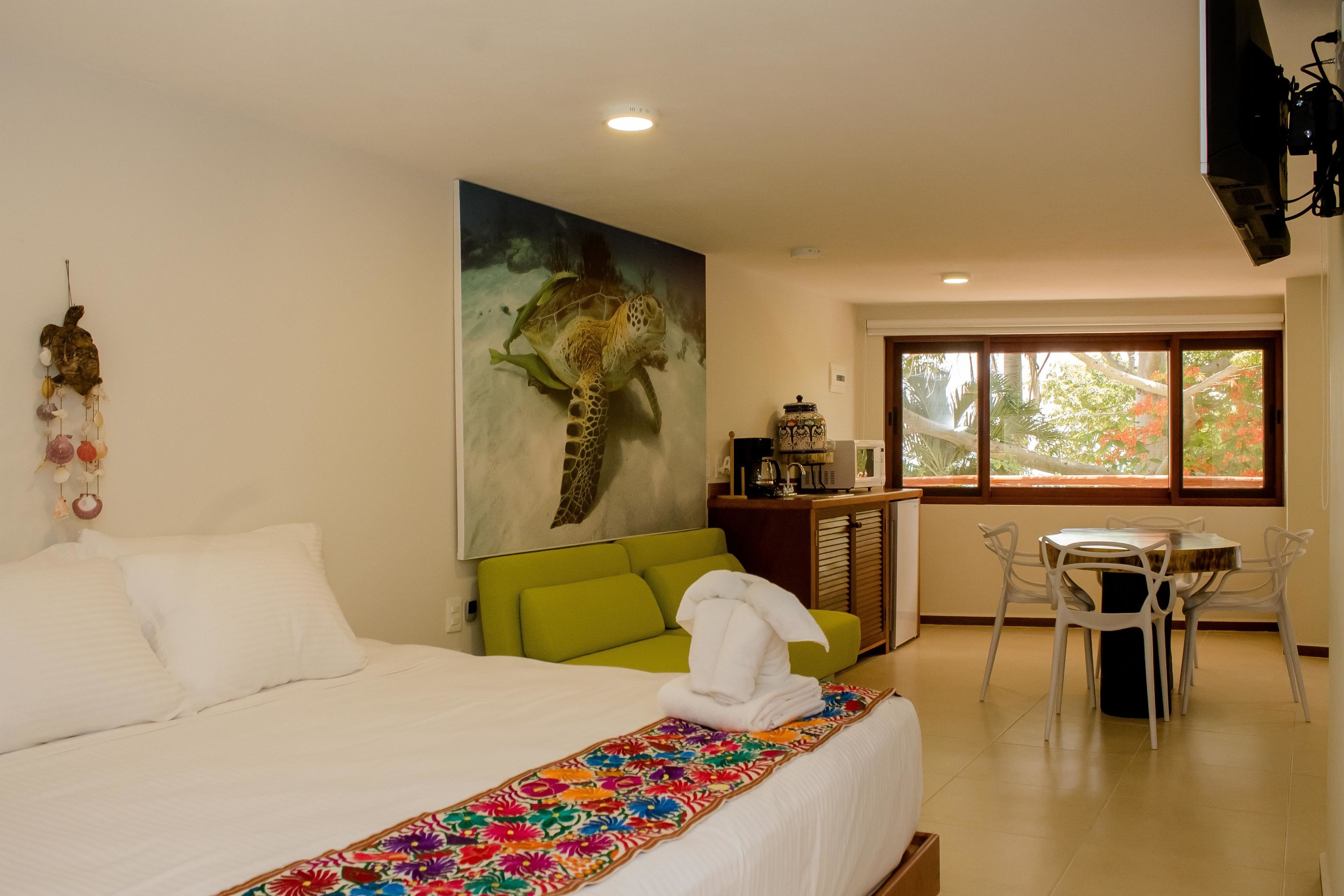 Una habitación de hotel con cama, un sofá verde, una mesa de comedor con sillas y un cuadro grande de una tortuga marina en la pared.