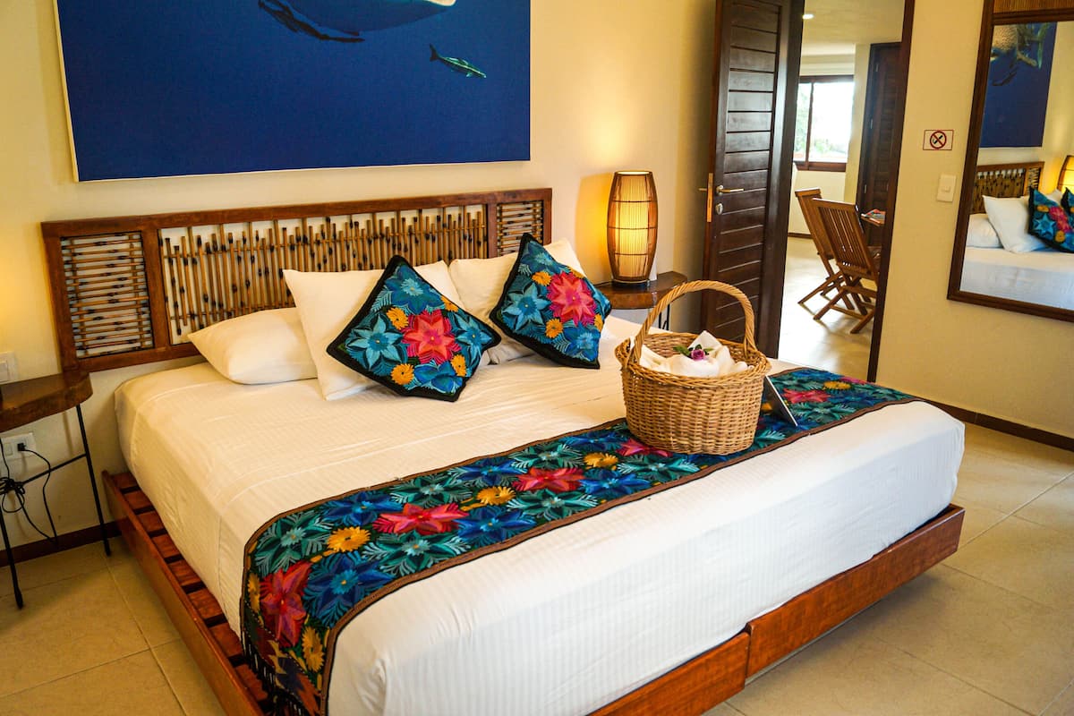 Una cama con cabecera de madera, dos almohadas, una colcha colorida y una cesta de mimbre al lado.