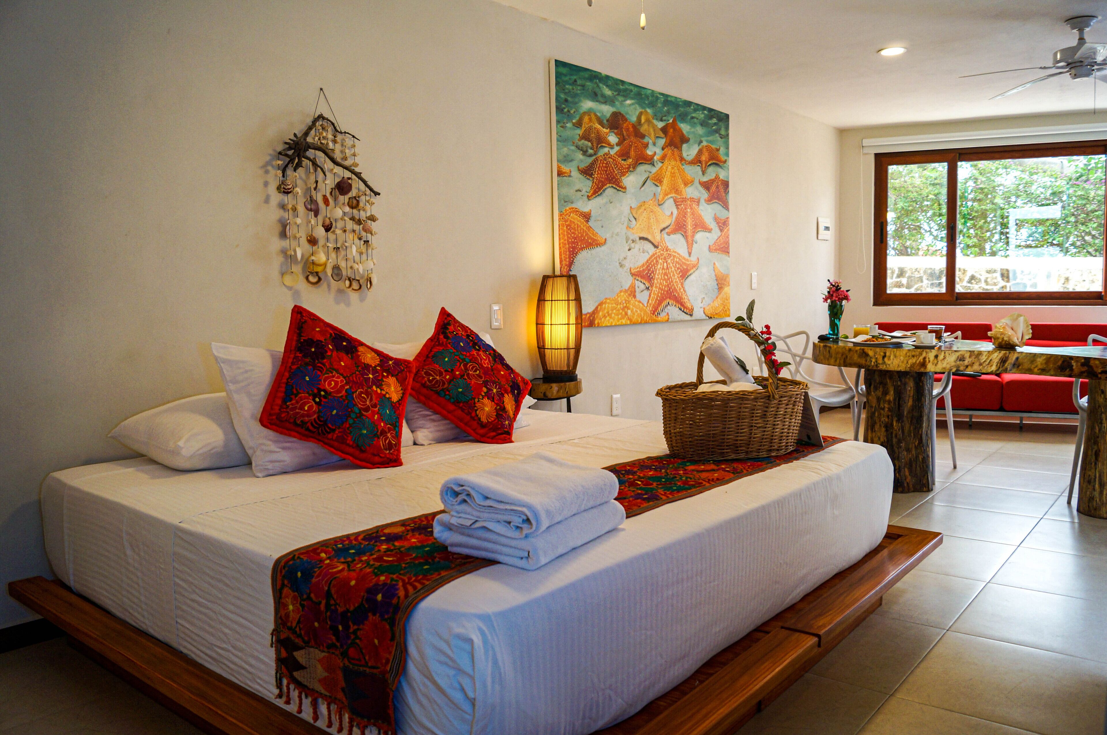 A neatly made bed with a decorative headboard, a wall painting, a table with a basket, and a red bench in a bright room with a large window.