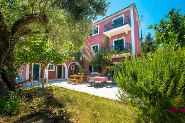 Ein zweistöckiges Gebäude mit roter Fassade, einem Balkon im oberen Geschoss und einer Terrasse mit Tisch und Stühlen.