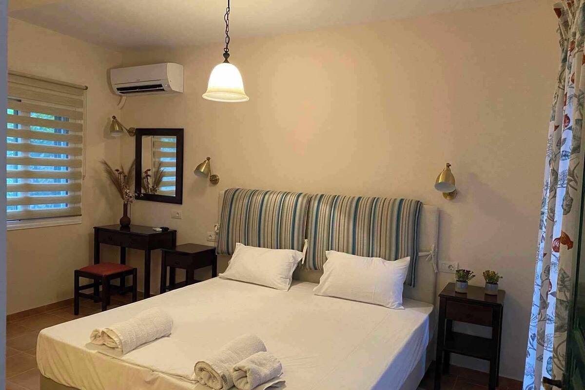 A neatly made bed with white linens and rolled towels, a bedside table with a lamp, a striped headboard, and an air conditioning unit on the wall.