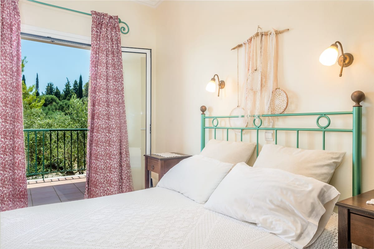 A bedroom with a bed, a nightstand, a window with curtains, and a view of greenery outside.
