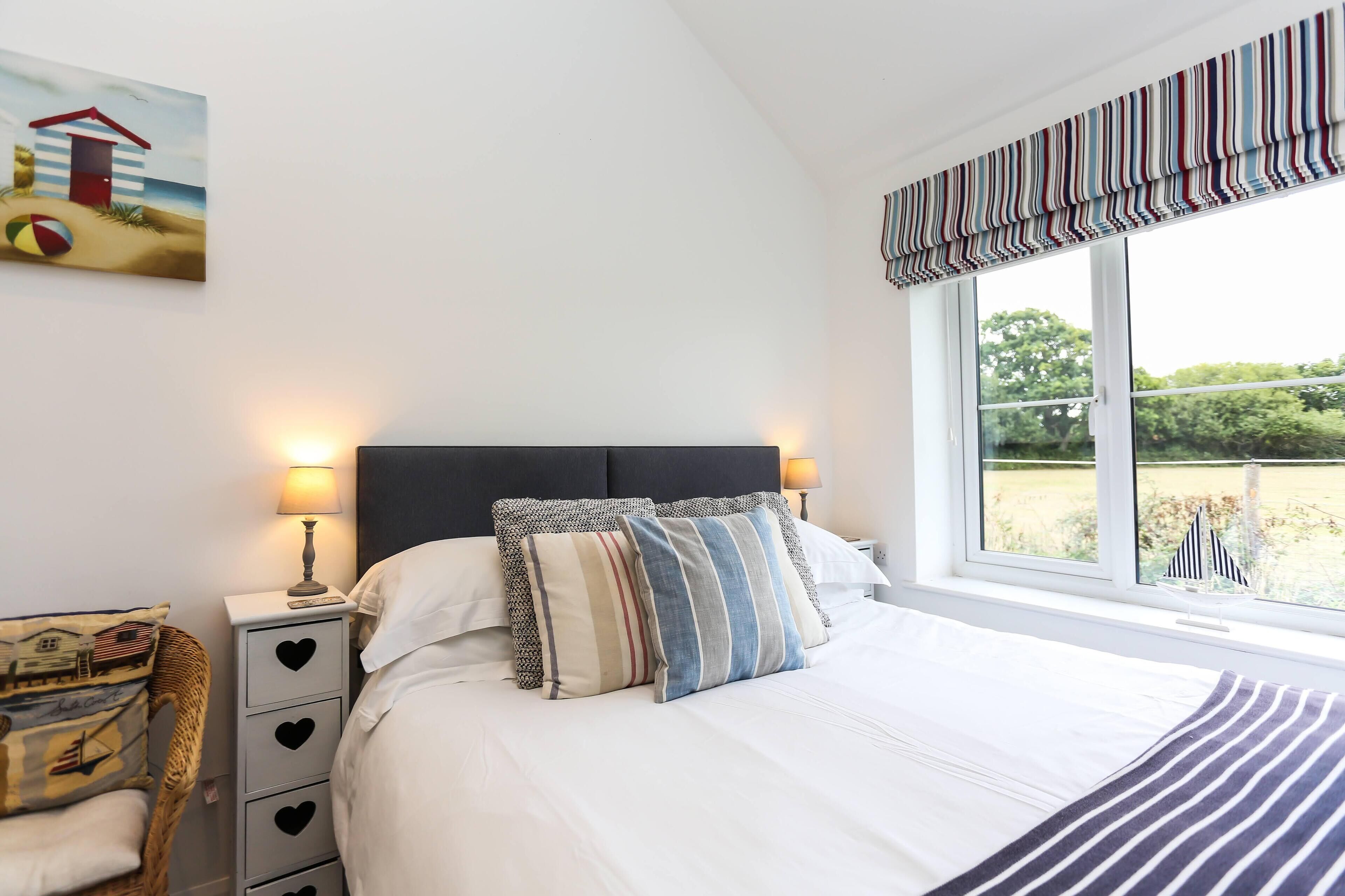 A neatly made bed with striped pillows, a bedside table with a lamp, a heart-shaped drawer, and a window with a view of the outdoors.