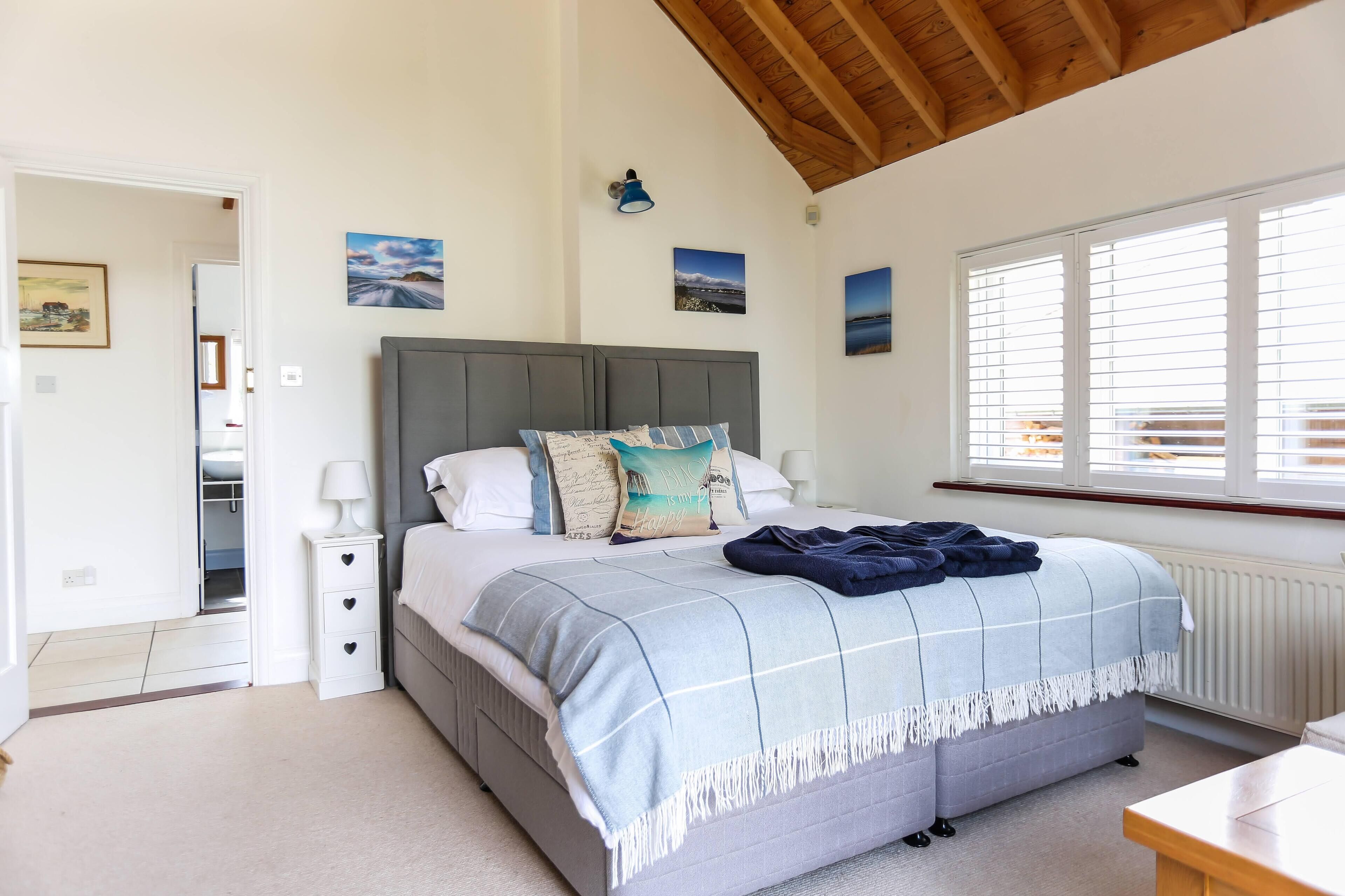 A bedroom with a grey upholstered bed, a bedside table with a lamp, a window with blinds, and a view of the outside.