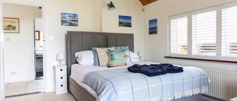 A bedroom with a grey upholstered bed, a bedside table with a lamp, a window with blinds, and a view of the outside.