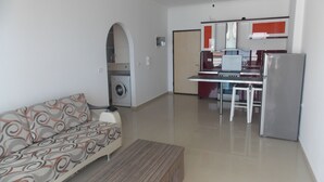A living room with a sofa, a coffee table, a washing machine, a kitchen with a refrigerator, and a dining area. - Belvedere Apartaments 2 (Sarandë)