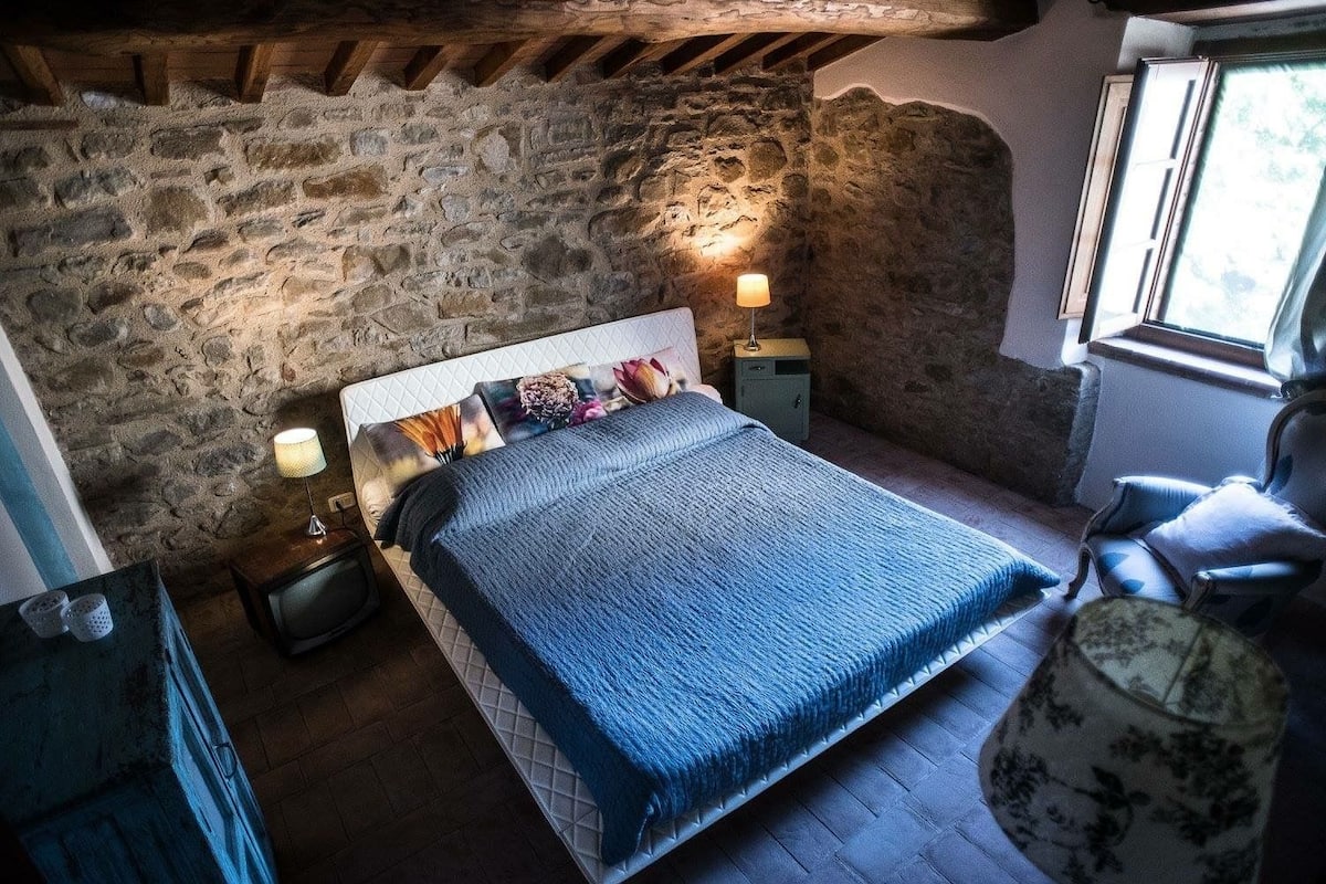 A bedroom with a stone wall, a bed with a blue blanket, a nightstand with a lamp, and a chair.