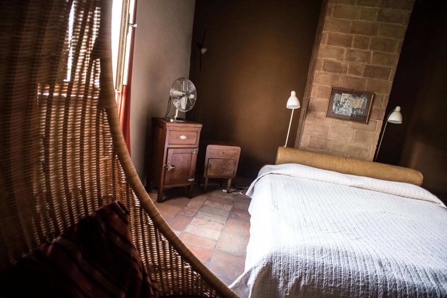 A bedroom with a bed, a wooden nightstand, a wicker chair, a small table, and a brick wall.