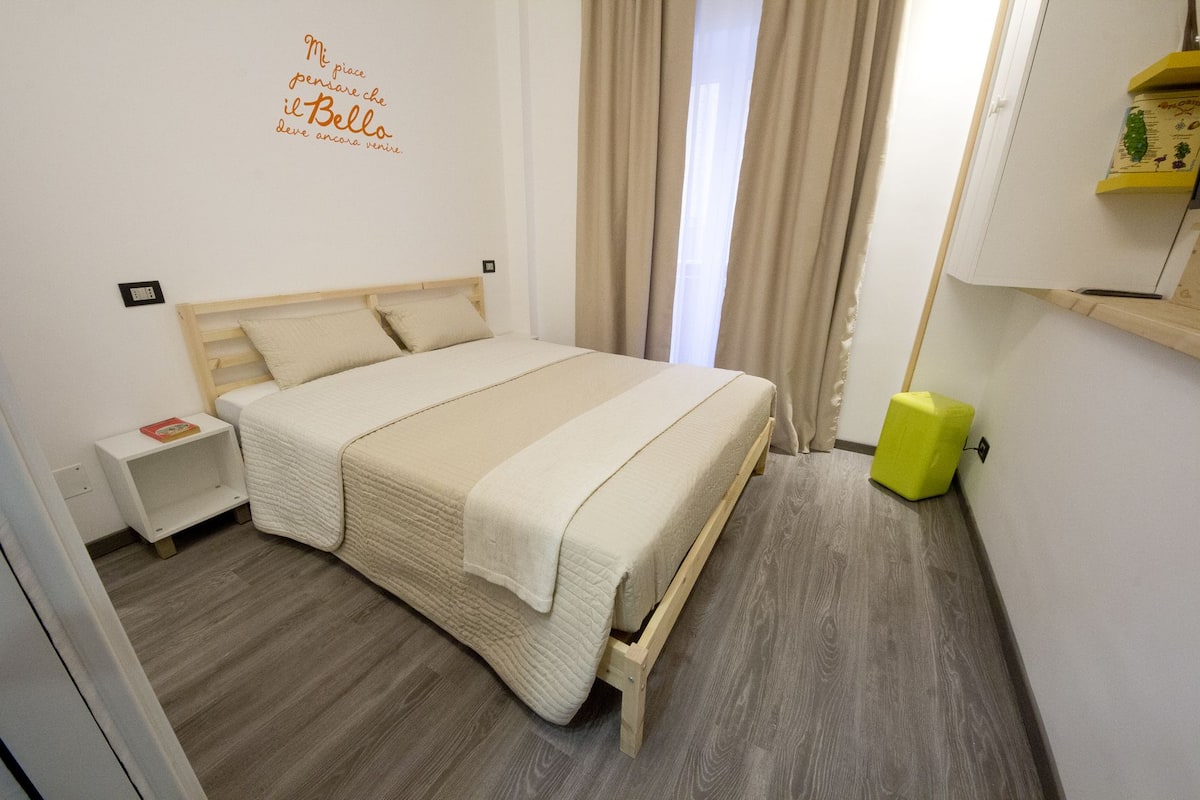 Cama bem arrumada num quarto de hotel, com cabeceira de madeira, mesa de cabeceira com um livro e uma pequena prateleira com uma planta.