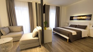 Una habitación de hotel moderna con sofá, cama, una mesita con una planta y un cuadro de paisaje urbano en la pared.