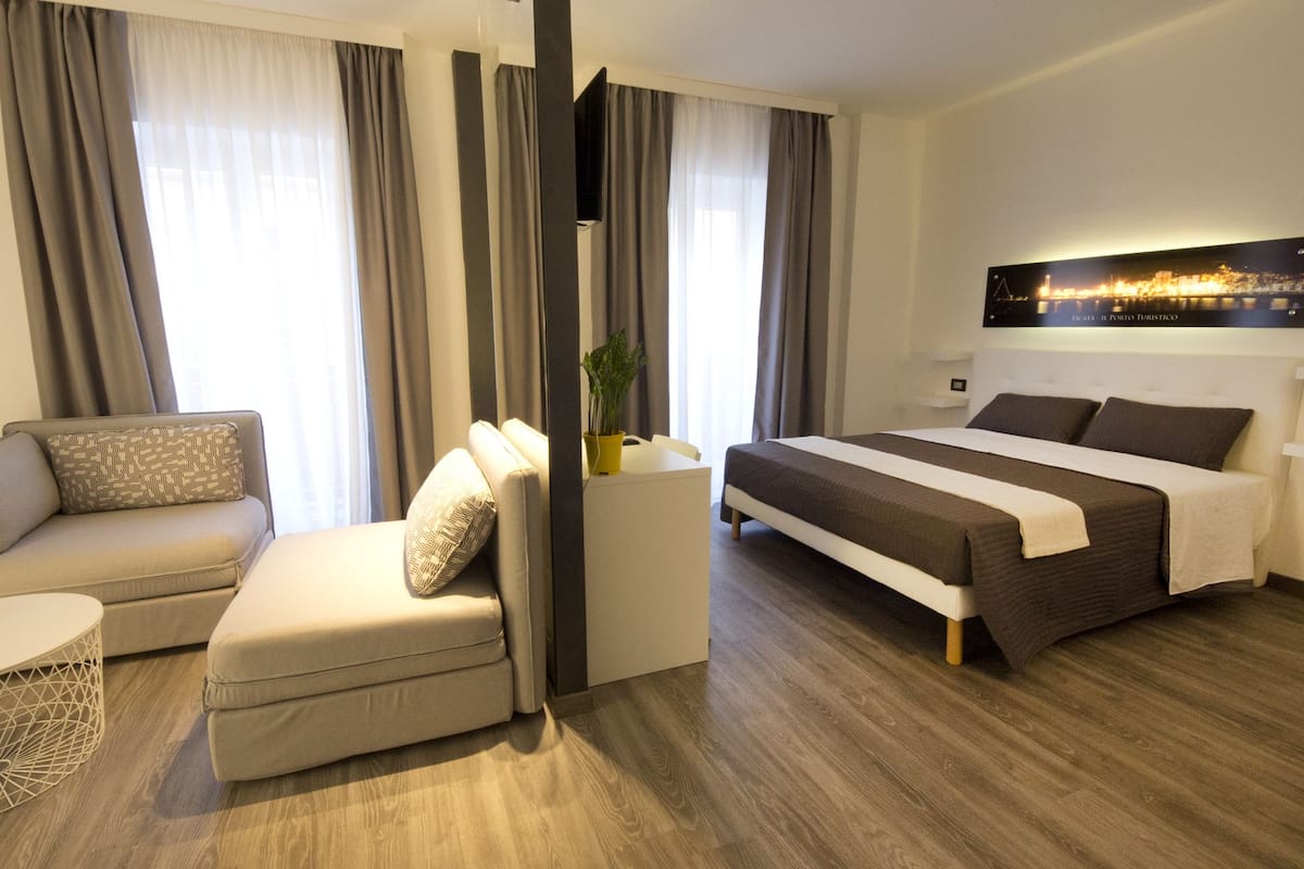 Quarto de hotel moderno com sofá, cama, uma pequena mesa com planta e uma pintura de paisagem urbana na parede.