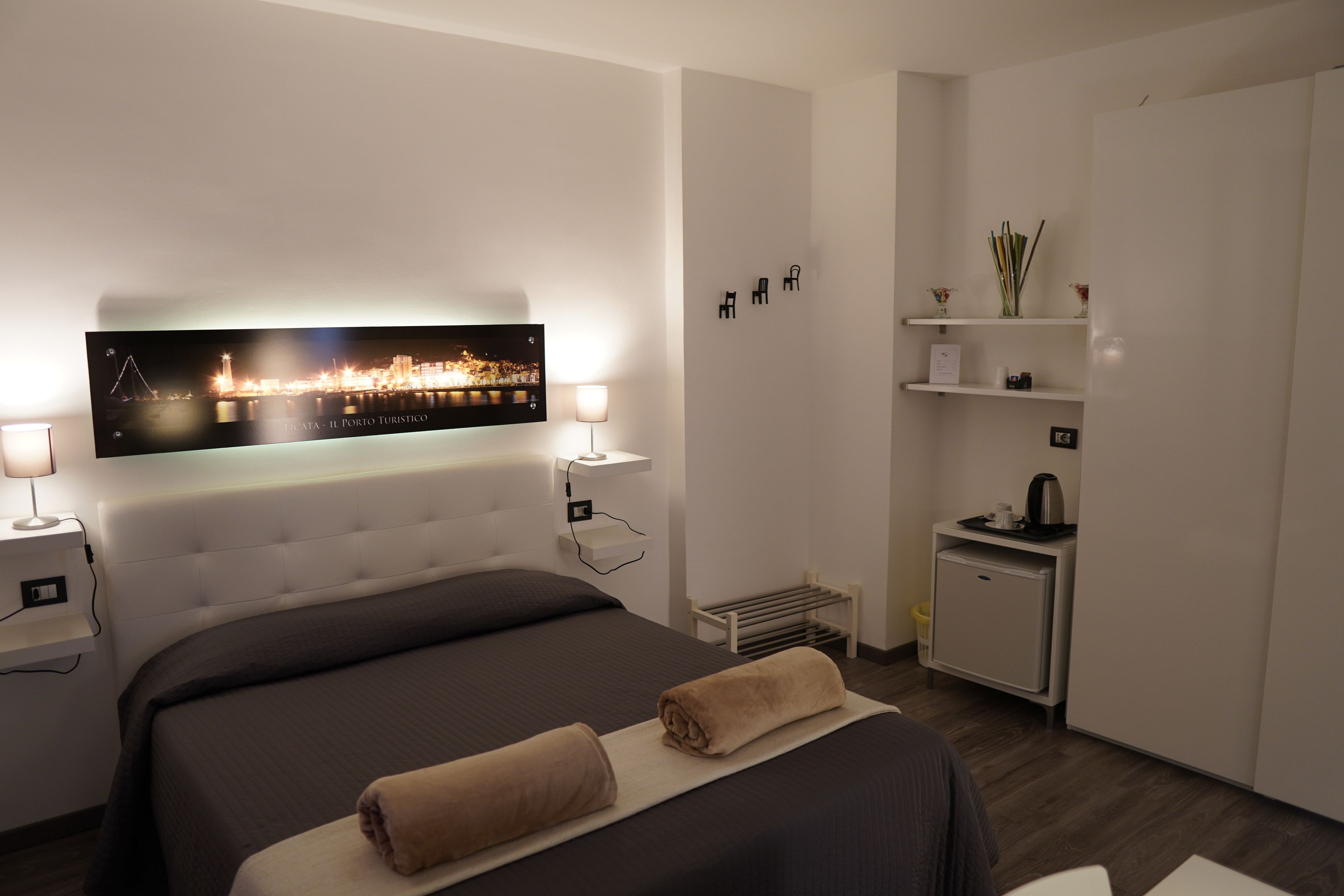 A modern hotel room with a large bed, bedside lamps, a flat-screen TV displaying a cityscape, a small kitchenette, and a walk-in closet.