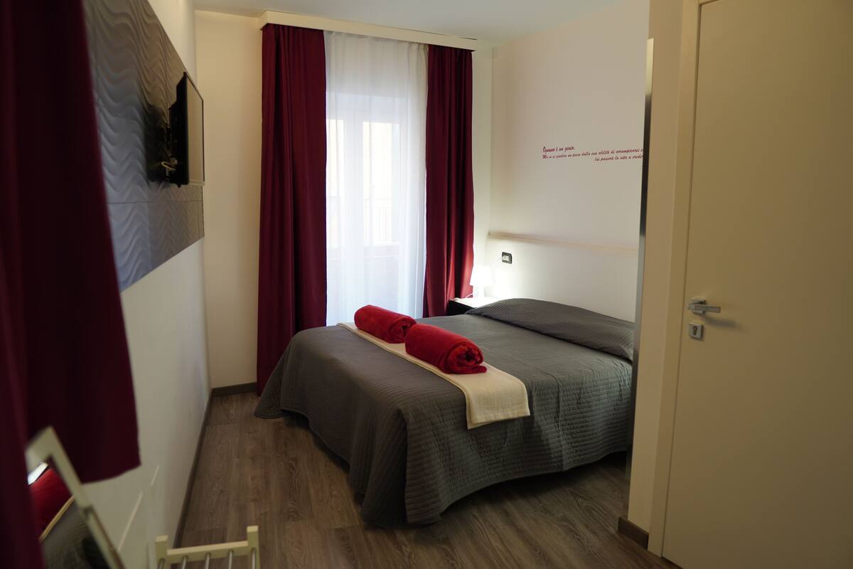 Quarto de hotel com uma cama, televisão fixada na parede e uma janela com cortinas.