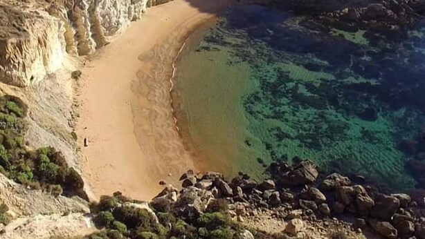 Paisaje costero con playa arenosa, aguas azules cristalinas, acantilados rocosos y un edificio pequeño a la izquierda.