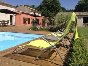 Ett trädäck med gula solstolar vid en pool, omgivet av grönska och en röd byggnad.