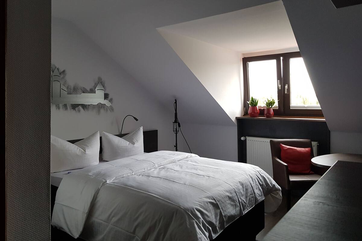 A bedroom with a sloped ceiling, a bed with white bedding, a desk with a red pillow, and a window with plants.