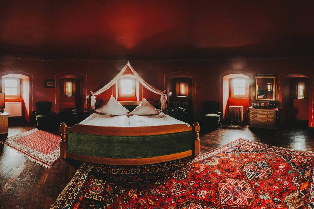 A large, ornate bed with a canopy in a dimly lit room with red walls and multiple windows.