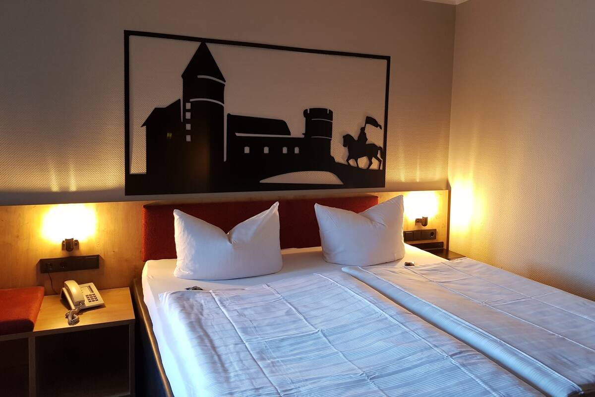 A hotel room with a bed, two white pillows, a nightstand with a telephone, and a wall-mounted artwork depicting a cityscape.