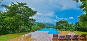 Una terraza de madera con piscina, rodeada de abundante vegetación y montañas al fondo.