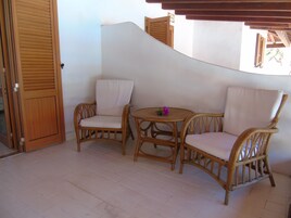 Esquina de una habitaci ón con dos sillones de mimbre, una mesita redonda y una puerta de madera, además de una pared blanca.