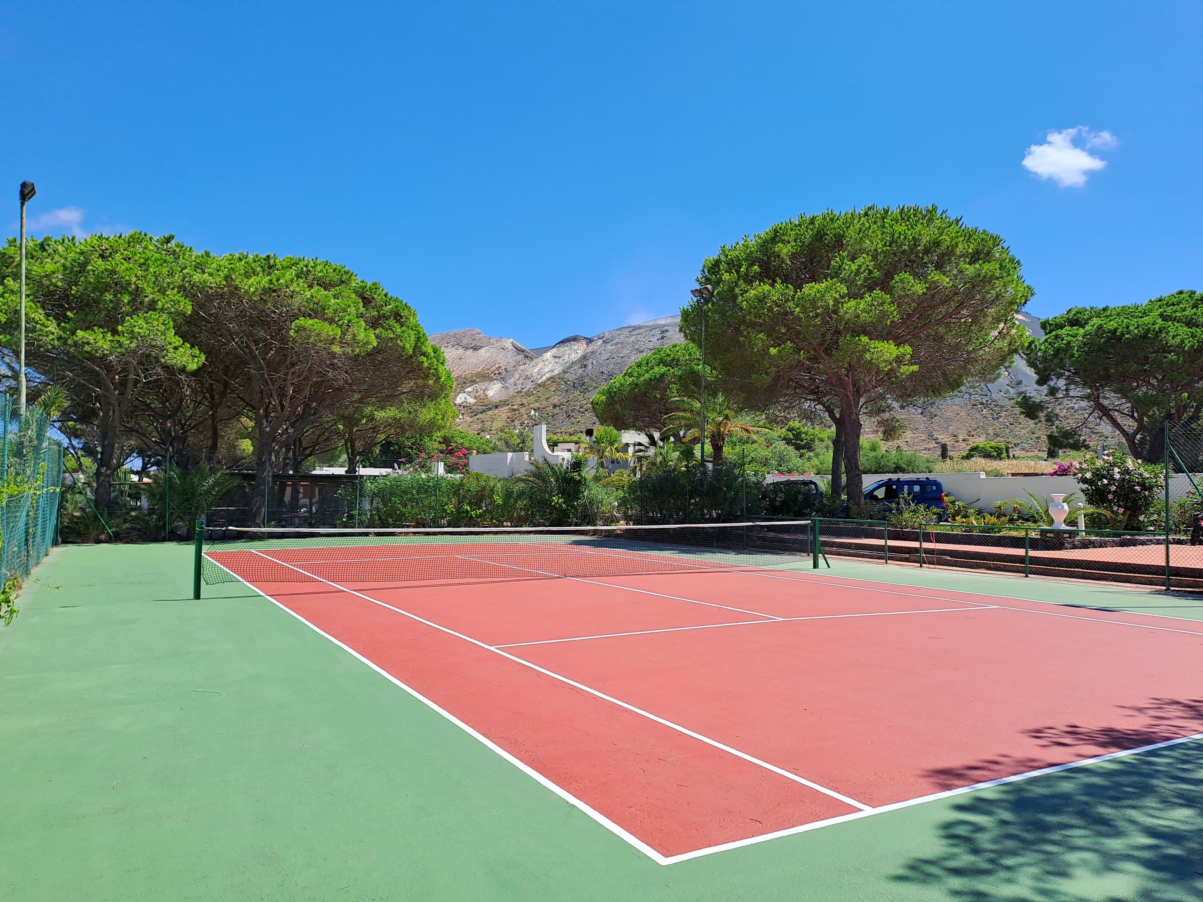 A tennis court with a green and red surface, surrounded by trees and a mountainous backdrop.