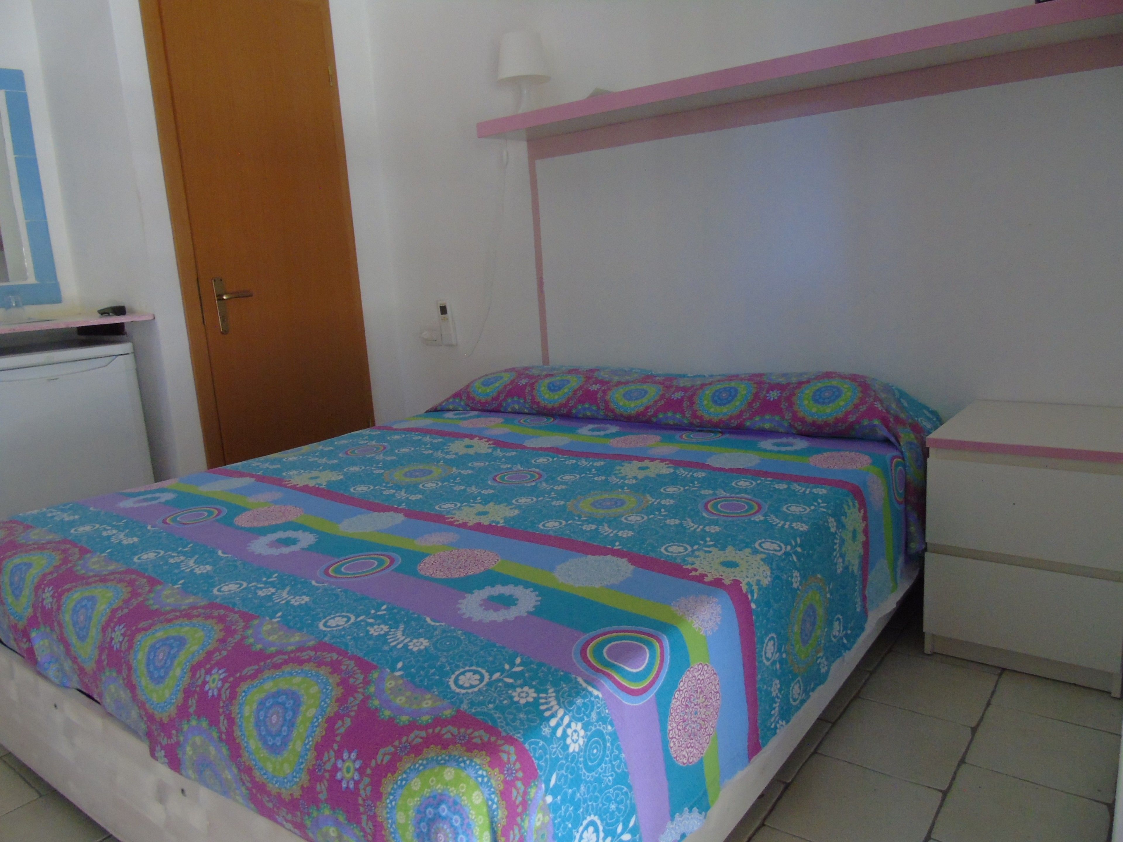 A bedroom with a bed, a dresser, a door, and a small white appliance.