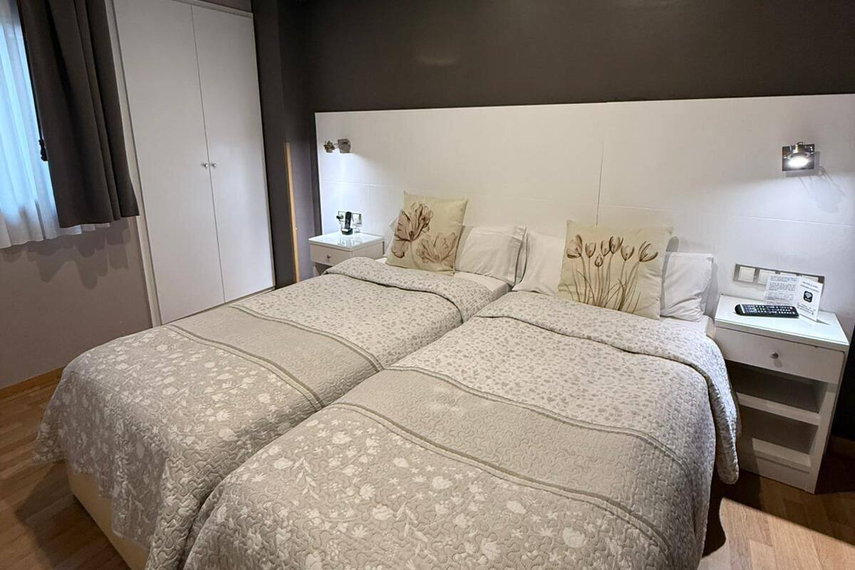 Una cama doble con una colcha de motivos florales, una mesita de noche con una lámpara y una mesita auxiliar con un mando a distancia.