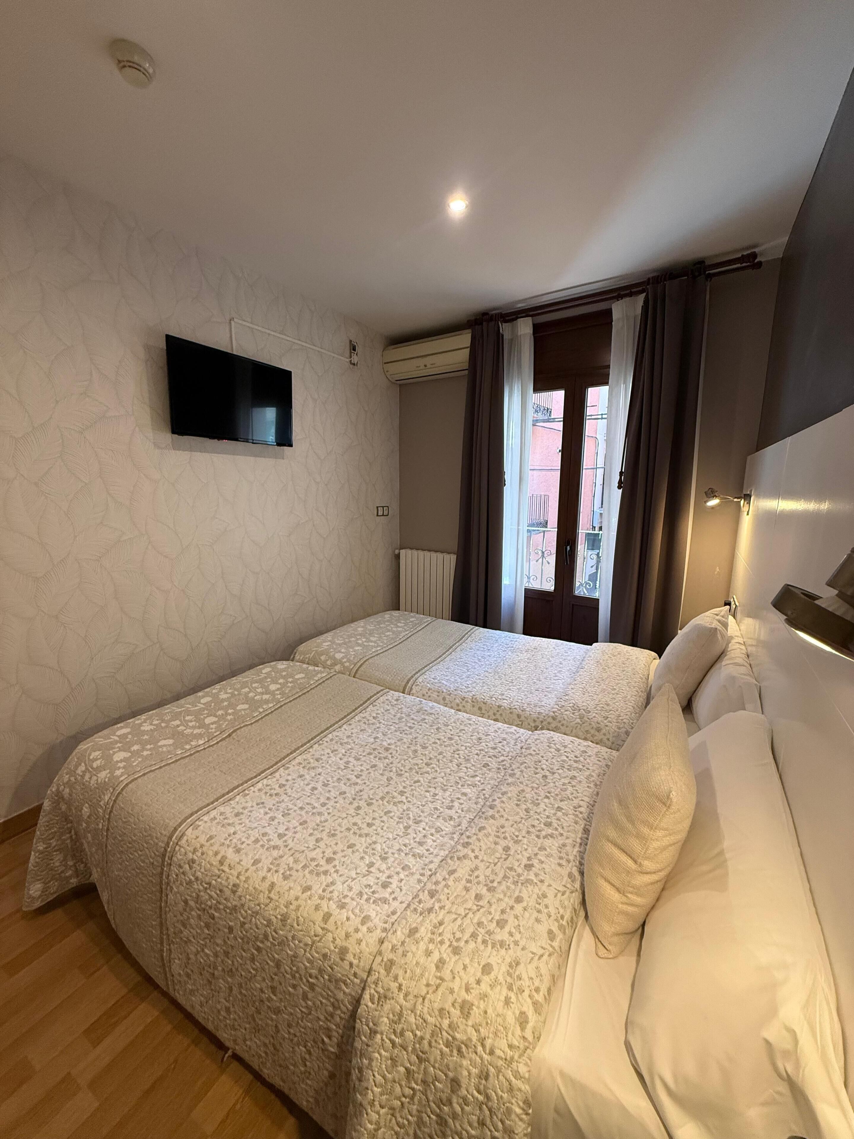 Habitación de hotel con dos camas, un televisor de pantalla plana montado en la pared, un equipo de aire acondicionado y una ventana con cortinas.