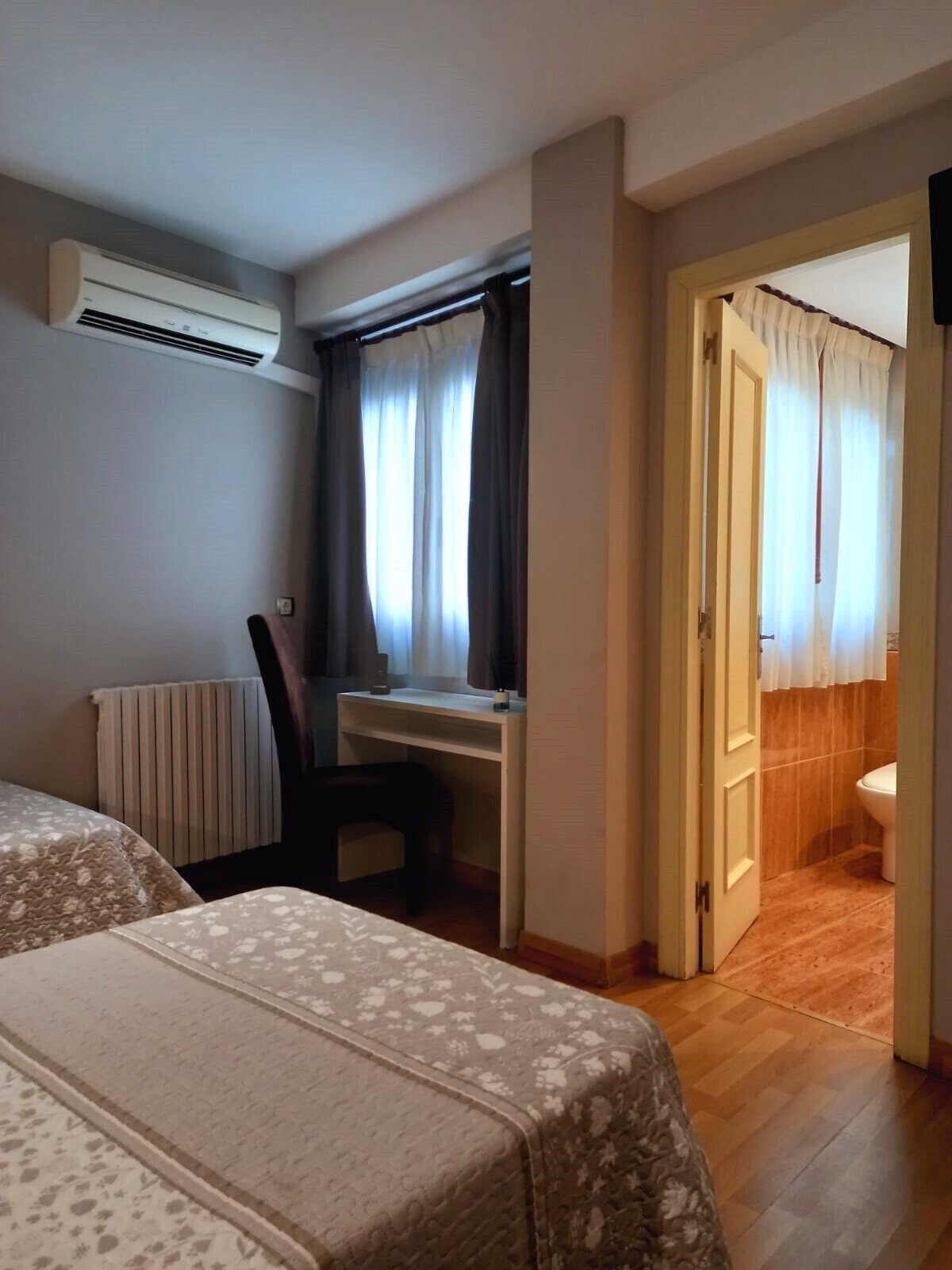 Ein Hotelzimmer mit zwei Betten, einem Schreibtisch, einem Stuhl, einer Klimaanlage und Blick ins Bad durch eine offene Tür.