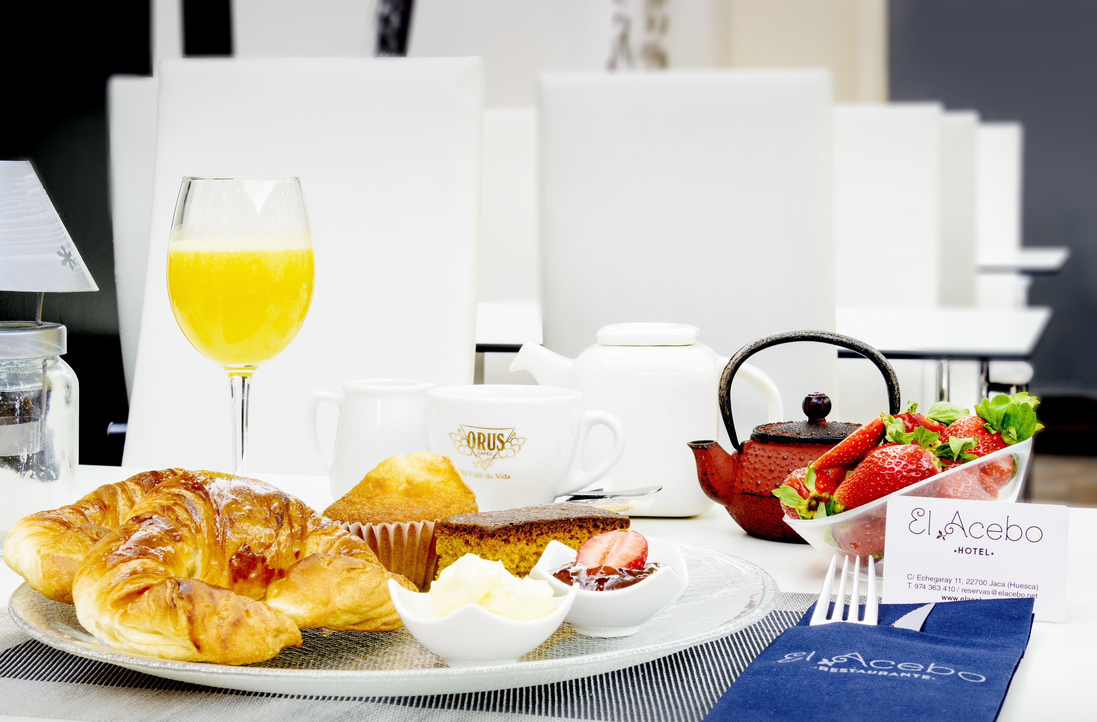 Desayuno con cruasanes, un vaso de jugo de naranja, un muffin, una taza, una tetera, fresas y un letrero del Hotel El Acebo.