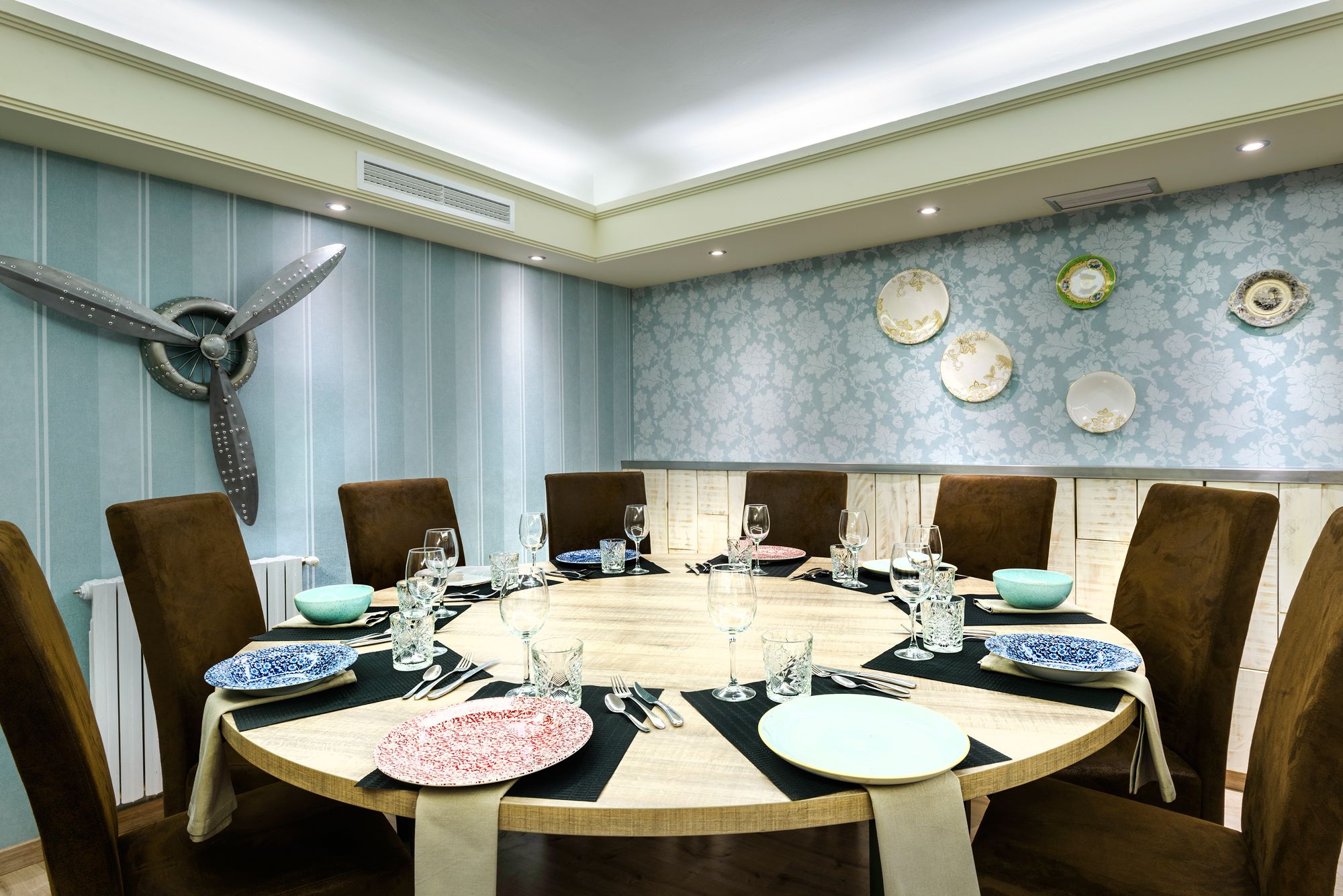Una mesa redonda de comedor con seis sillas, rodeada de un papel tapiz azul y blanco con diseño y platos decorativos en la pared.