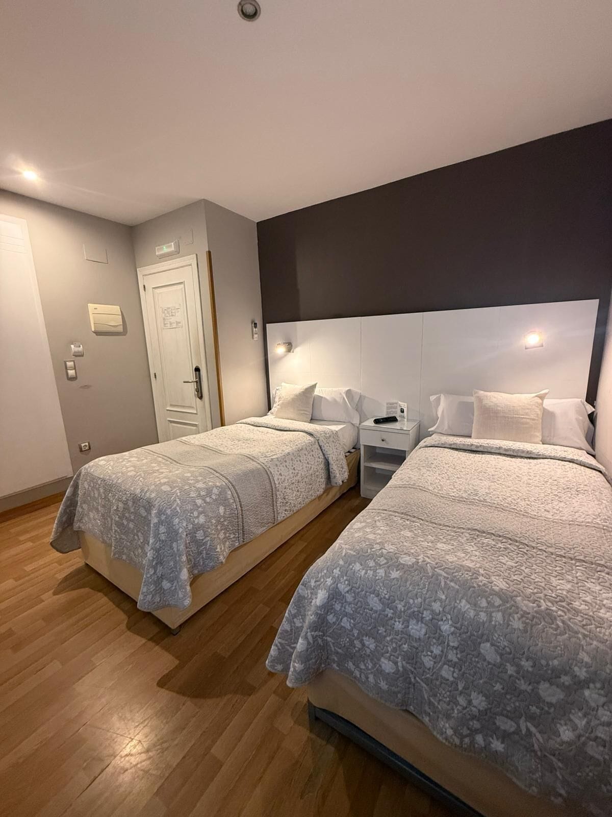 Una habitación de hotel con dos camas, una mesita de noche, una puerta blanca y un termostato en la pared.