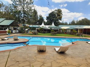 Piscina con sillones de playa y sombrillas, rodeada de césped y árboles.
