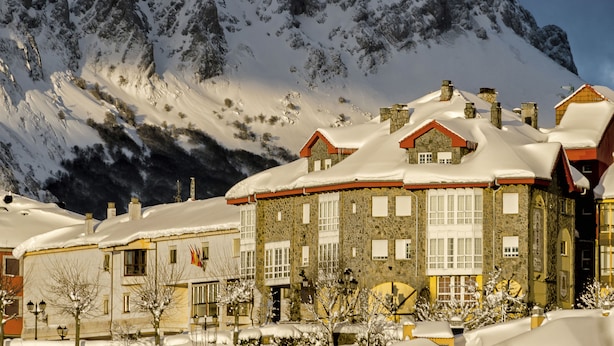 Un edificio cubierto de nieve con muros de piedra y varias chimeneas, frente a montañas con cimas nevadas.