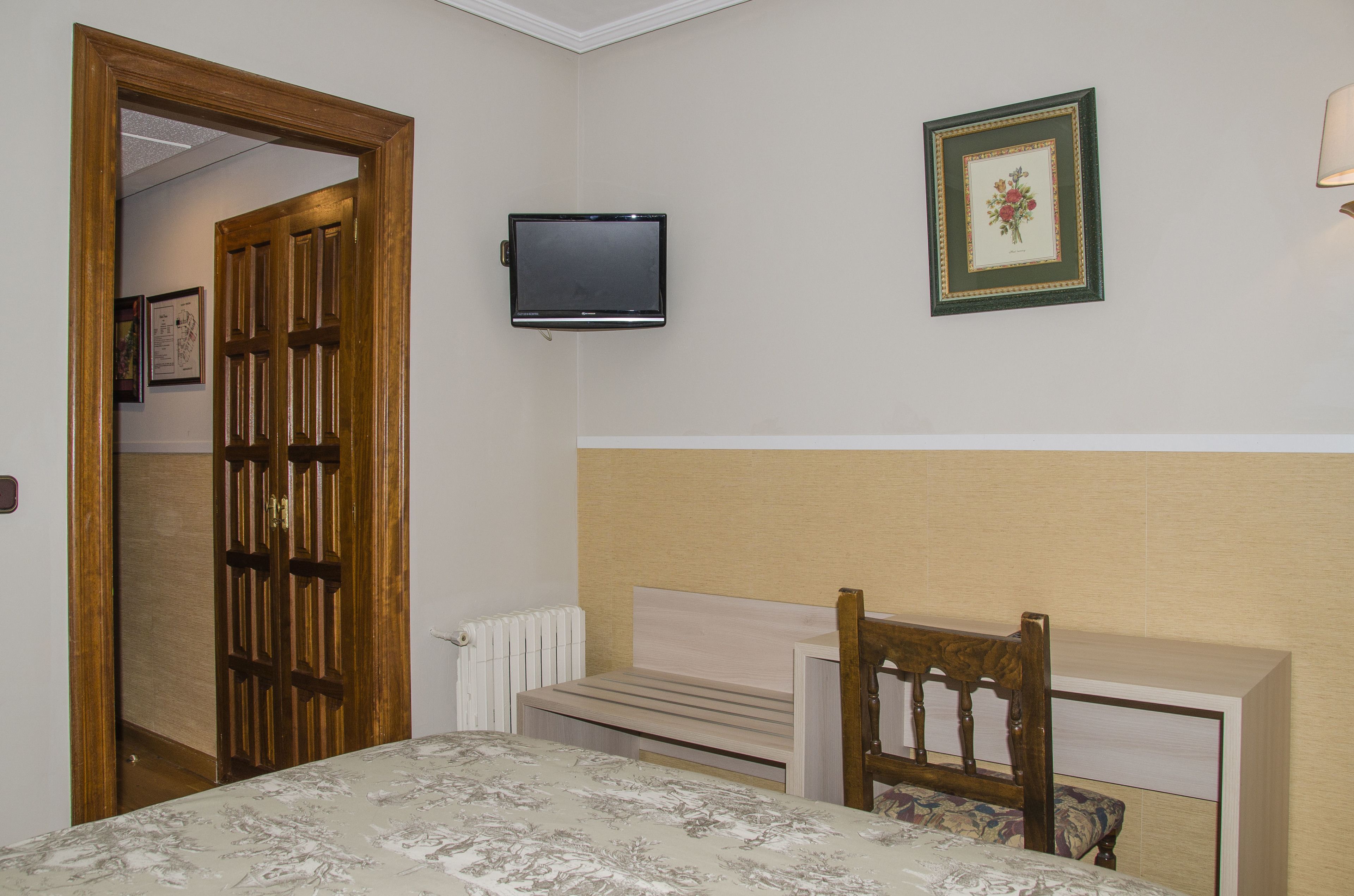 Un dormitorio con cama, mesita de noche, silla, televisor montado en la pared y puerta que da a otra habitación.