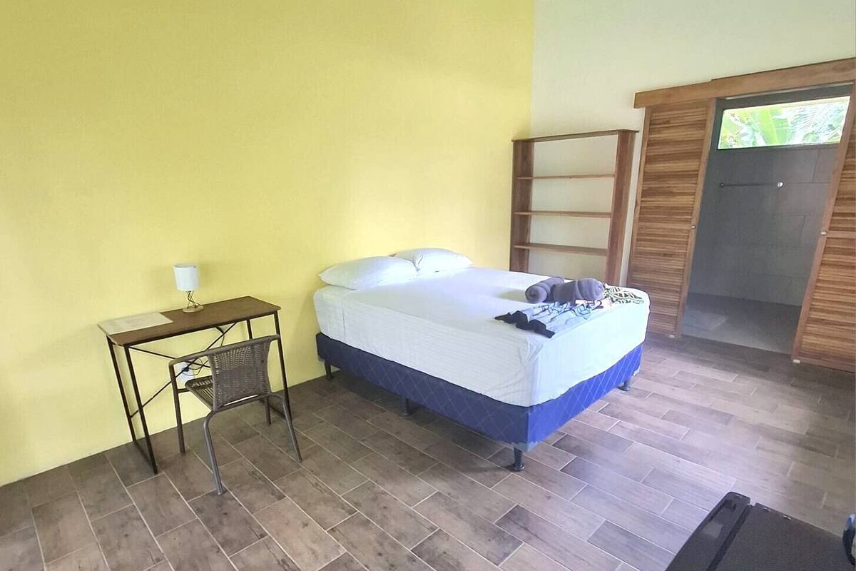 Quarto com cama, uma mesinha com abajur, ventoinha no teto e porta de madeira.