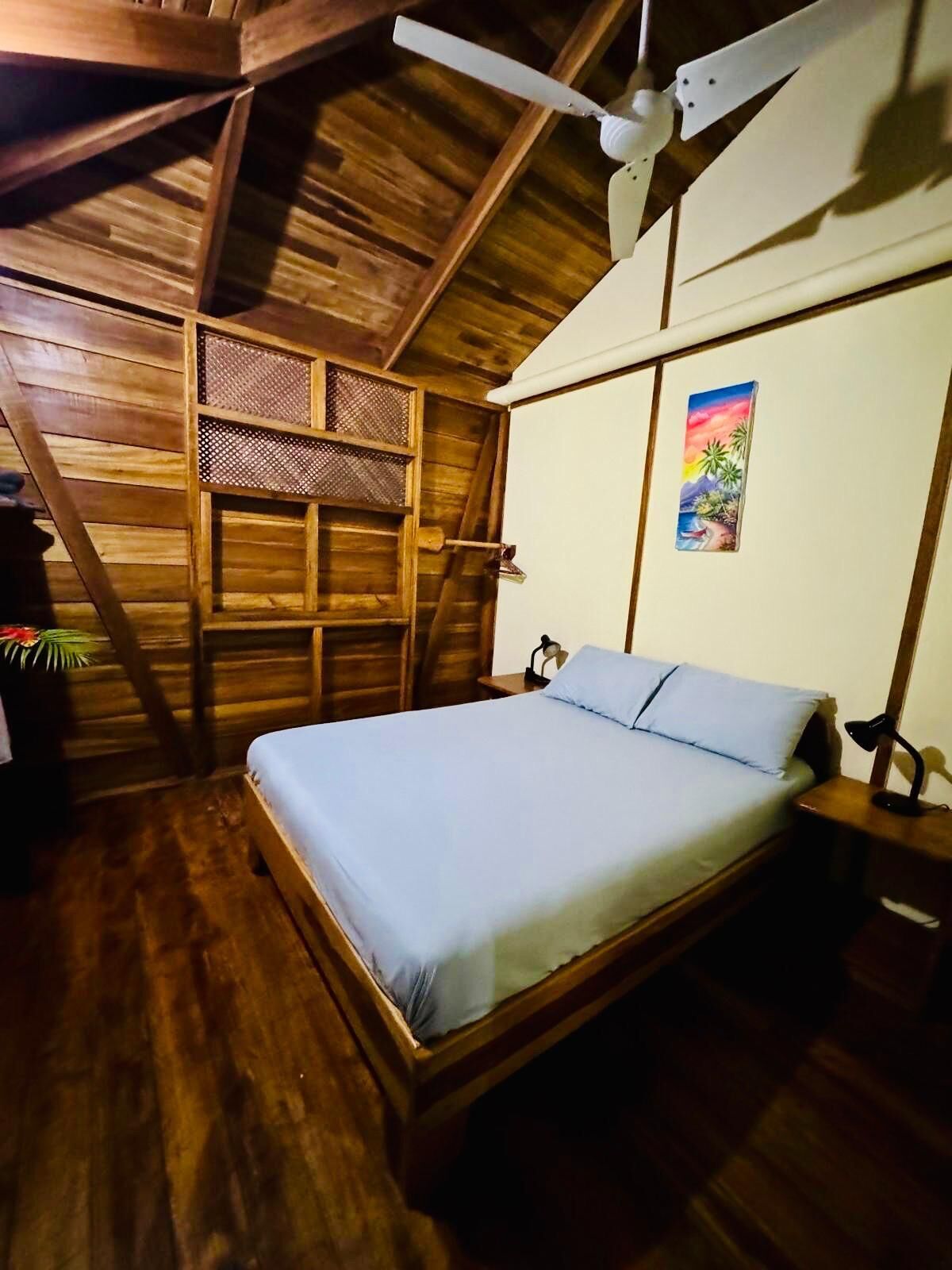 Une chambre avec un plafond en bois, un lit avec un matelas bleu, une table de chevet avec une lampe et un tableau accroché au mur.