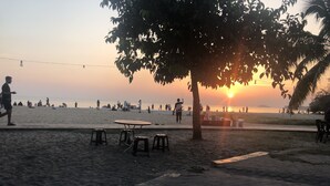 Cena de praia com uma mesa e banquetas sob uma árvore, pessoas caminhando e um pôr do sol ao fundo.